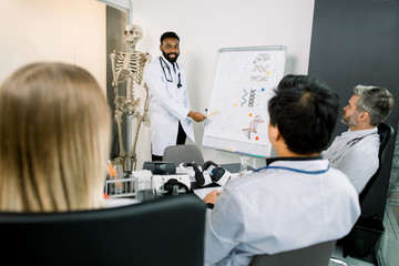 Obraz premium Handsome young African man doctor scientist wearing white coat standing near flipchart and giving presentation about biochemical molecules, new medicines to multiethnic group of his colleagues