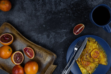 Two crepe suzette on a plate, golden toasted with slices of red orange and in syrup on a dark background. In a wooden tray are fruits - the ingredients of the filling. Flat lay with a copy place.