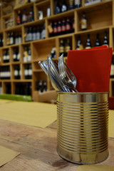Long wooden table set by placeholder red napkins and cutlery in a wine shop in a Tuscan restaurant. In the background a wall of wines on display with wooden shelves with bokeh effect.