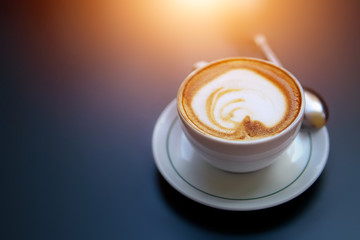 Aromatic cappuccino with latte foam in white cup on low key dark background. Copy space. Sun light effect. Coffee break concept, early wake up, arabica coffee bar