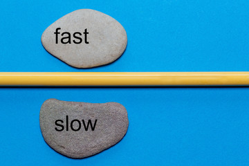 The words fast and slow are written on natural smooth stones separated by a yellow pencil. The...