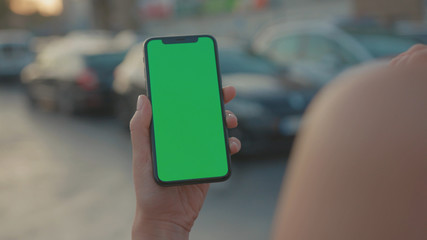 Lviv, Ukraine - May 19, 2018: Young woman hitchhiking on night highway holding and using modern mock-up vertical smartphone greenscreen outdoors. Close-up.