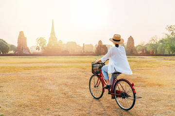 Woman riding a bicycle with front basket  near Ayutthaya historical park in Thailand, a beautiful early morning time with bright sunrise light. Careless Asian vacation concept image. - obrazy, fototapety, plakaty