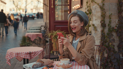 Happy young woman with red lipstick hat wearing stylish coat typing on phone during sunny day sitting in cafe on breakfast outdoors smile city fashion online stylish slow motion