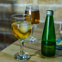 On the dining table is a glass of cold sparkling apple juice with an ice cube and a green bottle. Small foamy beer on a blurred background. Selective focus, Closeup view. Square frame