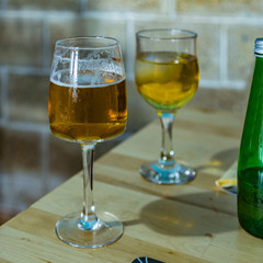 On the dining table is a glass of small foamy beer and a green bottle. Cold gglass of sparkling apple juice with an ice cube on a blurred background. Selective focus, Closeup view. Square frame