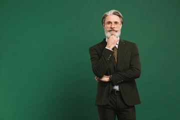 A bearded middle-aged teacher in khaki suit holding a beard and thinking on a green background with...