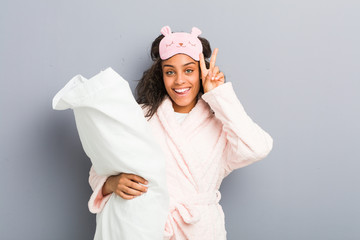 Young african american woman wearing a pajamas and a sleep mask holding a pillow showing victory...