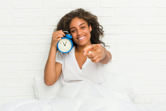Young african american woman sitting on the bed holding an alarm clock cheerful smiles pointing to front.