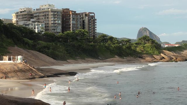 Steady shot of people enjoying the ocean waves of Devils beach seen from the Arpoador rock in Ipanema with the Sugarloaf mountain in the background