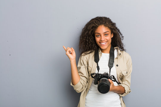 Young african american photographer woman holding a camera smiling cheerfully pointing with forefinger away.