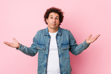 Curly mature man wearing a denim jacket against pink background doubting and shrugging shoulders in...