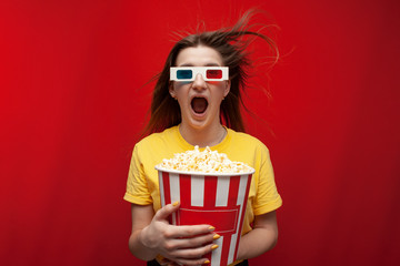 funny young girl student watching a movie in 3D glasses and screaming on a red background, she eats popcorn and is delighted