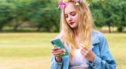 Cheerful young woman blonde hair wearing flowers around reading message or social media with smartphone in her hands while walking with glass orange juice in hand at park. Technology internet concept.
