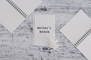 piece of paper on wooden desk with Writer's Block text surrounded by blank notepads and with with scrunched paper ball next to it