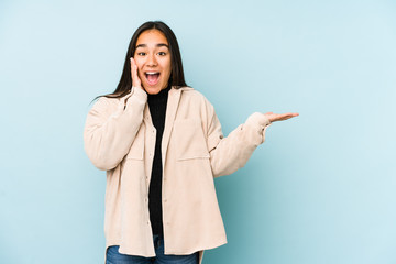 Young woman isolated on a blue background holds copy space on a palm, keep hand over cheek. Amazed and delighted.