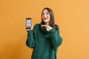 Young woman with menstrual calendar on screen of mobile phone on color background
