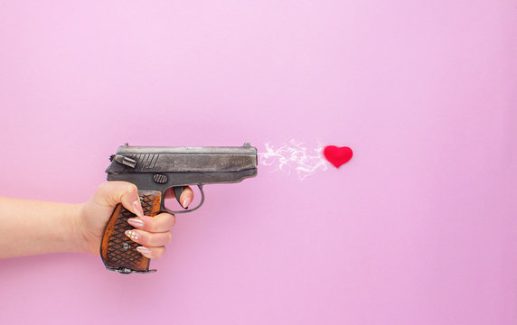 A gun firing a heart in a female hand on a pink background. Love concept