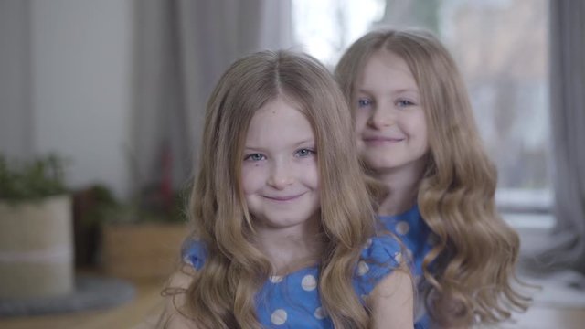 Two little pretty Caucasian twins looking at each other and turning to camera smiling. Happy cute children posing at home. Lifestyle, happiness, leisure, unity. Camera moving away.