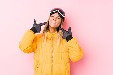 Young caucasian woman wearing a ski clothes in a pink background smiles, pointing fingers at mouth.