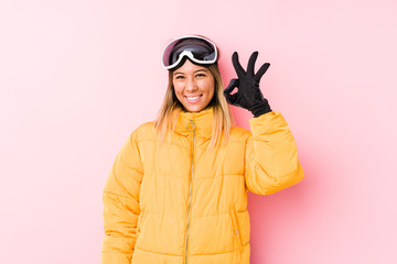 Young caucasian woman wearing a ski clothes in a pink background winks an eye and holds an okay gesture with hand.
