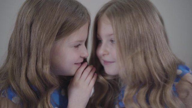 Close-up of little pretty Caucasian girl whispering on twin sister's ear and showing hush gesture. Joyful twins keeping secrets of each other. Lifestyle, unity, happiness.