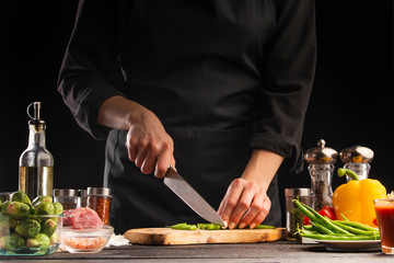 The chef cuts green beans on the background of vegetable ingredients, on a black background. Cooking salad. Healthy and wholesome food, cuisine and cooking, recipe book.