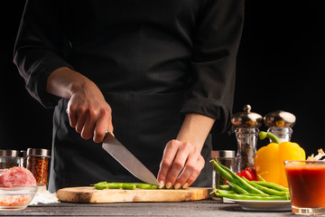 The chef cuts green beans on the background of vegetable ingredients, on a black background. Cooking salad. Healthy and wholesome food, cuisine and cooking, recipe book.