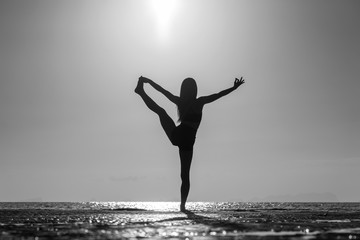 Fototapeta na wymiar Silhouette of woman standing at yoga pose on the tropical beach during sunset. Girl practicing yoga near sea water. Black and white