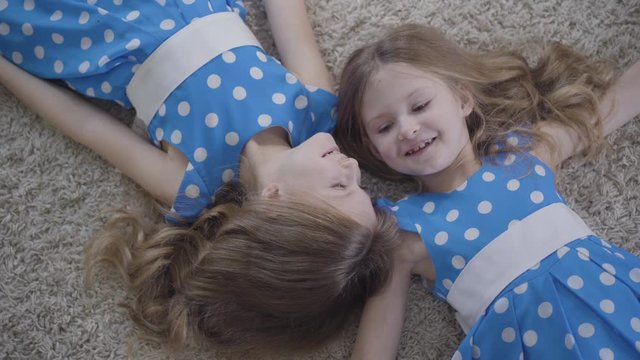 Top view of two pretty Caucasian twin sisters lying on soft carpet and smiling. Camera moving around faces of cute brunette girls resting at home. Leisure, lifestyle, unity.