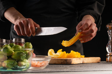 Chef slices sweet orange bell pepper on the background of vegetable ingredients, on a black background. Cooking salad. Healthy and wholesome food, cuisine and cooking, recipe book.