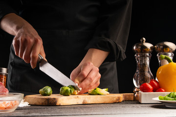 Chef slices Brussels sprouts on the background of vegetable ingredients, on a black background. Cooking salad. Healthy and wholesome food, cuisine and cooking, recipe book.