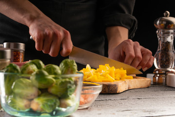 Chef slices sweet orange bell pepper on the background of vegetable ingredients, on a black background. Cooking salad. Healthy and wholesome food, cuisine and cooking, recipe book.