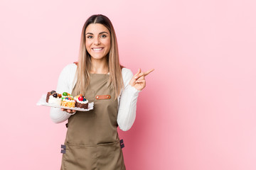 Young baker woman holding sweets smiling and pointing aside, showing something at blank space.