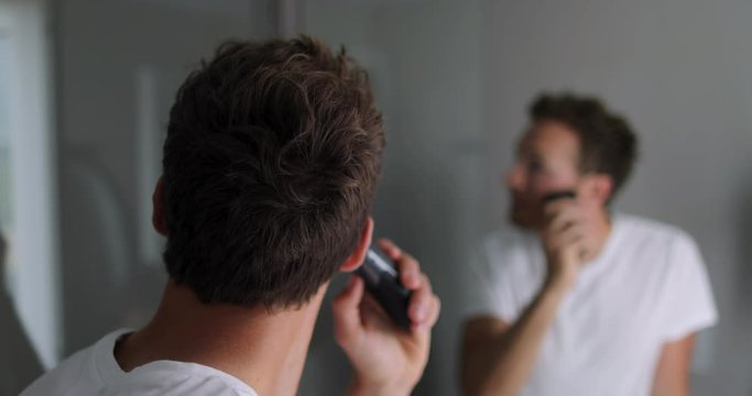 Man shaving beard using electric trimmer shaver. Male beauty grooming concept. Home lifestyle young person looking at bathroom mirror trimming hair on neck. 4K (UHD) 59.94 FPS SLOW MOTION.