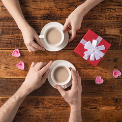 Valentine's Day celebration concept. A nice gift for your loved one. Hands of man and woman with coffee mugs on a wooden table background. Copy space. Flat lay. Close-up.