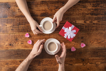 Valentine's Day celebration concept. A nice gift for your loved one. Hands of man and woman with coffee mugs on a wooden table background. Copy space. Flat lay. Close-up.