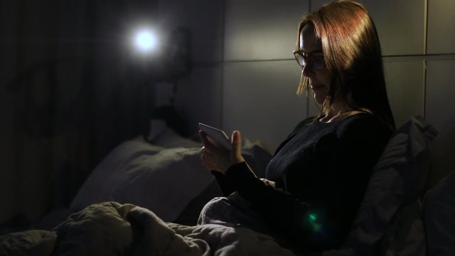 Adult woman puts on glasses and reads an e-reader device. Medium shot, dark night lighting.