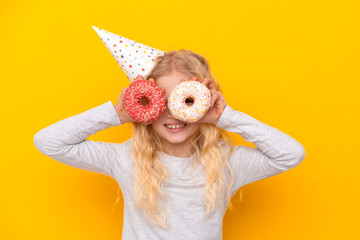 Crazy cheerful blonde girl in birthday hat smiling, having fun and looking through two donuts on her eyes. Sweets. Yellow studio background.