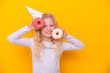 Crazy cheerful blonde girl in birthday hat smiling, having fun with two red donuts. Happy birthday and Sweets. Yellow studio background.