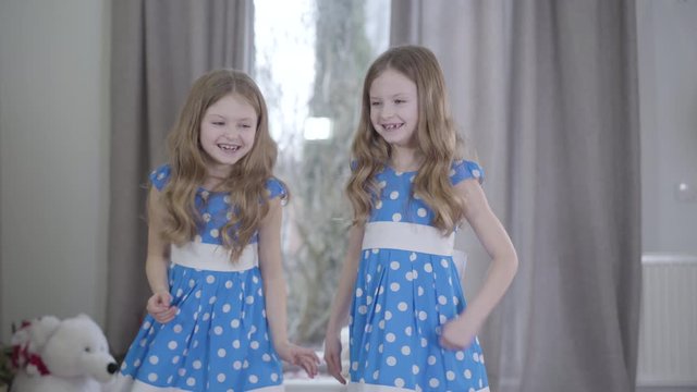 Middle shot of joyful twin sisters having fun indoors. Joyful brunette Caucasian girls in similar blue dotted dresses grimacing and smiling. Leisure, fun, lifestyle, family.