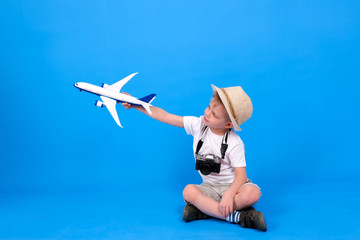 Full length blonde caucasian boy in summer hat and white clothes with camera playing toy airplane on blue. Family travel and tourism, summer holidays.