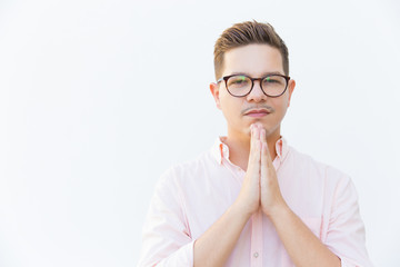 Handsome calm young man in eyeglasses praying. Young guy looking at camera with hands together in praying gesture. Hope and please concept