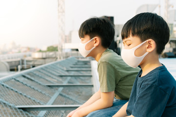 Two Asian boys sit on rooftop of building in city, look sad & desperate, wear medical face mask to protect from infection of virus, pandemic, influenza outbreak and epidemic of disease, wait for help.