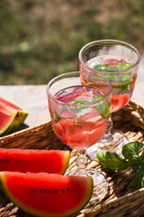 On a table in the garden in a basket are two glasses of lemonade and sliced of watermelon