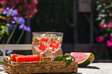 On a table in the garden in a basket are two glasses of lemonade and sliced of watermelon