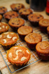 A lot of banana cupcakes on a cooling rack, some pieces have cashew nut or chocolate chip that on the wooden table