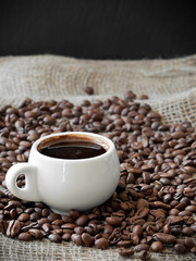 White cup with aromatic espresso coffee among coffee beans on burlap. Close-up, soft focus, copy space.