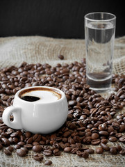 White cup with aromatic espresso coffee among coffee beans on burlap. Near a glass of water. Close-up, soft focus, copy space.