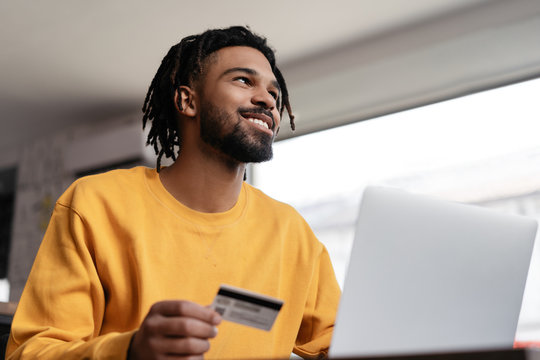 Successful Freelancer Receive Payment. Happy African American Man Holding Credit Card, Using Laptop Computer For Online Shopping.  Pensive Hipster Ordering Food Online, Booking Tickets And Smiling 